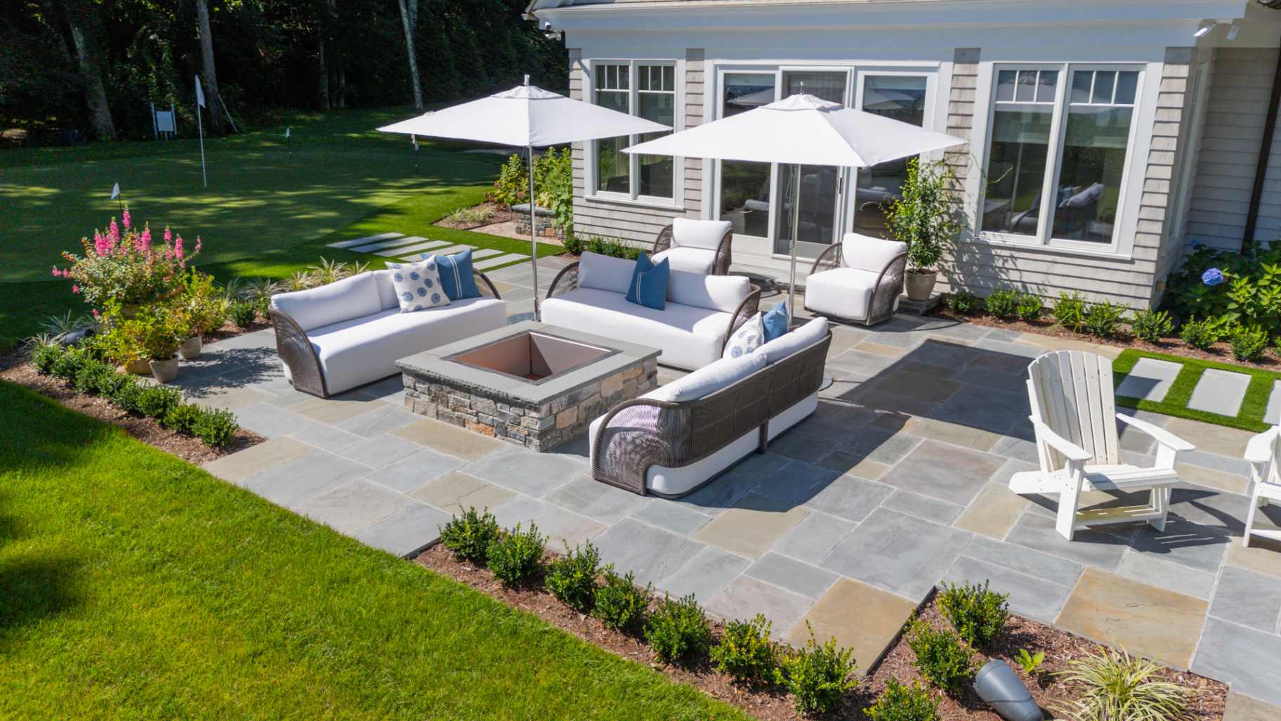 A bluestone patio with fire pit, lounge seating, and lush plantings beside a manicured lawn